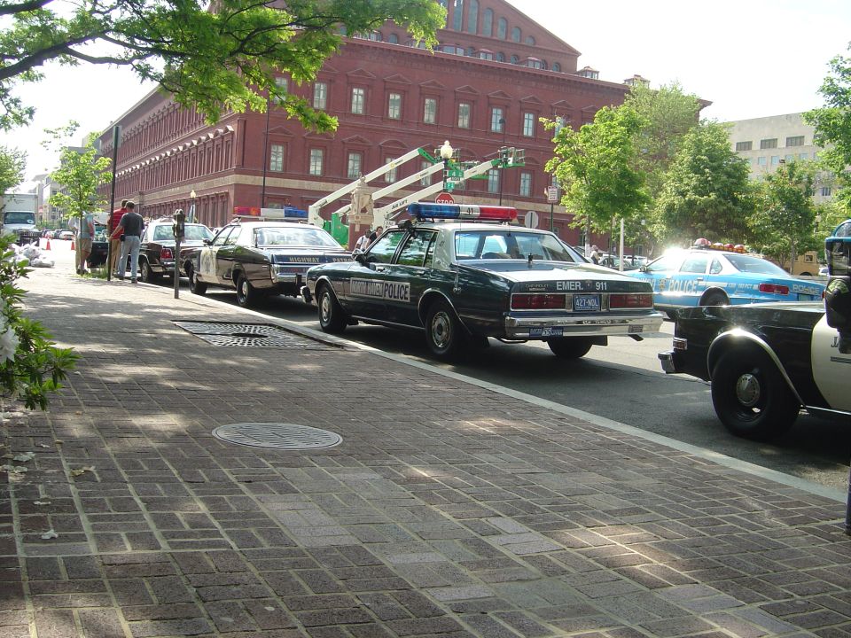 Police cars parked on a brick sidewalk, one dark green with "POLICE" text, another blue with light bar