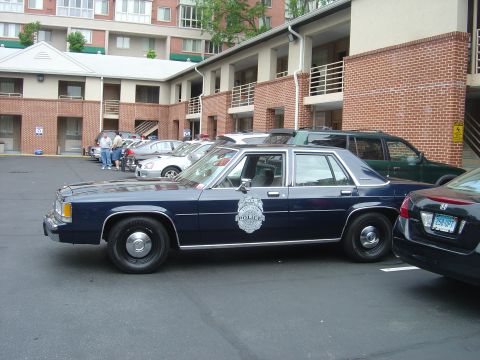 2001 Ford Crown Victoria - A dark blue police car is parked in a lot next to other vehicles, with a building in the background.