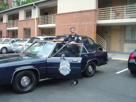 2001 Ford Crown Victoria - A police officer stands by a dark blue police car, with other vehicles parked in the background.
