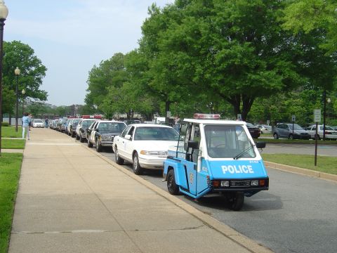 2001 Ford Crown Victoria - A line of police vehicles, including Crown Victorias and a three-wheeled patrol unit, parked on a street.