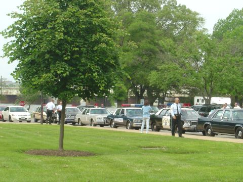 2001 Ford Crown Victoria - A line of police cars and civilians are gathered on a grassy area with trees in the background.