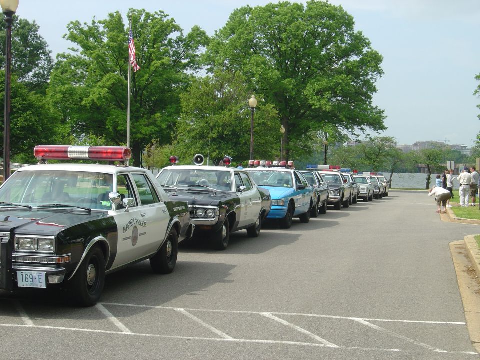 Line of police cars parked on asphalt, some with light bars. "Fairfax Police" text visible on one car