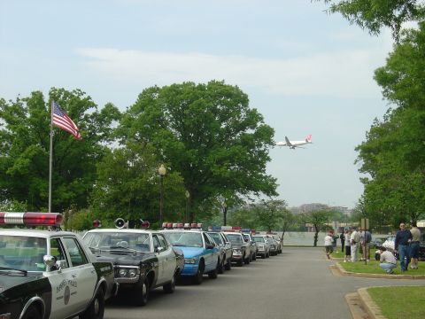 2001 Ford Crown Victoria - A row of police vehicles are parked on the side of the road, with an airplane in the sky above.