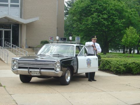 2001 Ford Crown Victoria - A police officer stands next to a vintage Plymouth police car parked in front of a building.