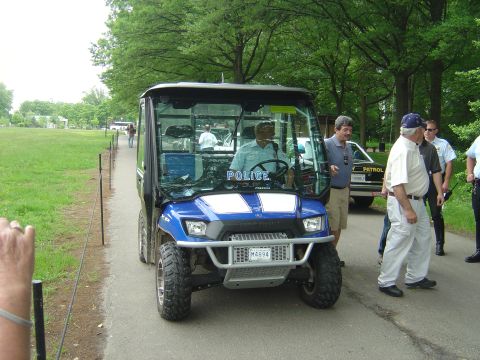 2001 Ford Crown Victoria - A blue and white utility vehicle with "POLICE" text is parked on a path, with several people standing nearby.