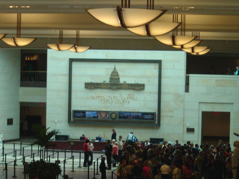 Interior of the United States Capitol Visitor Center with a large display of the Capitol building and text reading 'UNITED STATES CAPITOL VISITOR CENTER'. Many people are gathered