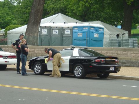 2001 Ford Crown Victoria - A police officer and two civilians stand near a black and white patrol car, with portable toilets and tents in the background.