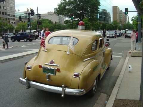 2001 Ford Crown Victoria - A vintage gold sedan with a light bar and flags drives down a city street.
