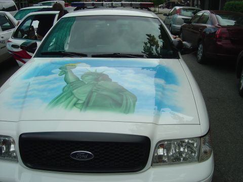 2001 Ford Crown Victoria - A white police car features an airbrushed Statue of Liberty on its hood, with other vehicles visible in the background.