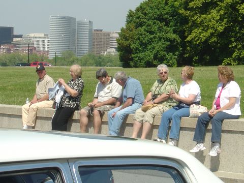 2001 Ford Crown Victoria - Foreground shows the hood and windshield of a white car, with a group of people sitting on a wall in the background against a city skyline.