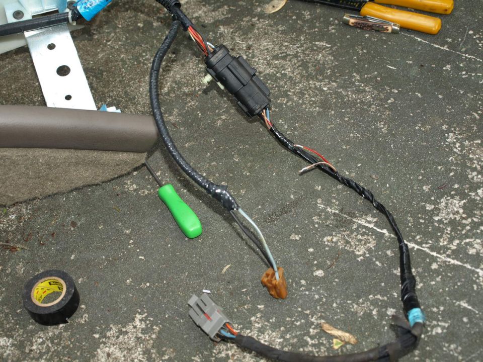 Car wiring harness with connectors, electrical tape, a green screwdriver, and a utility knife on a concrete surface