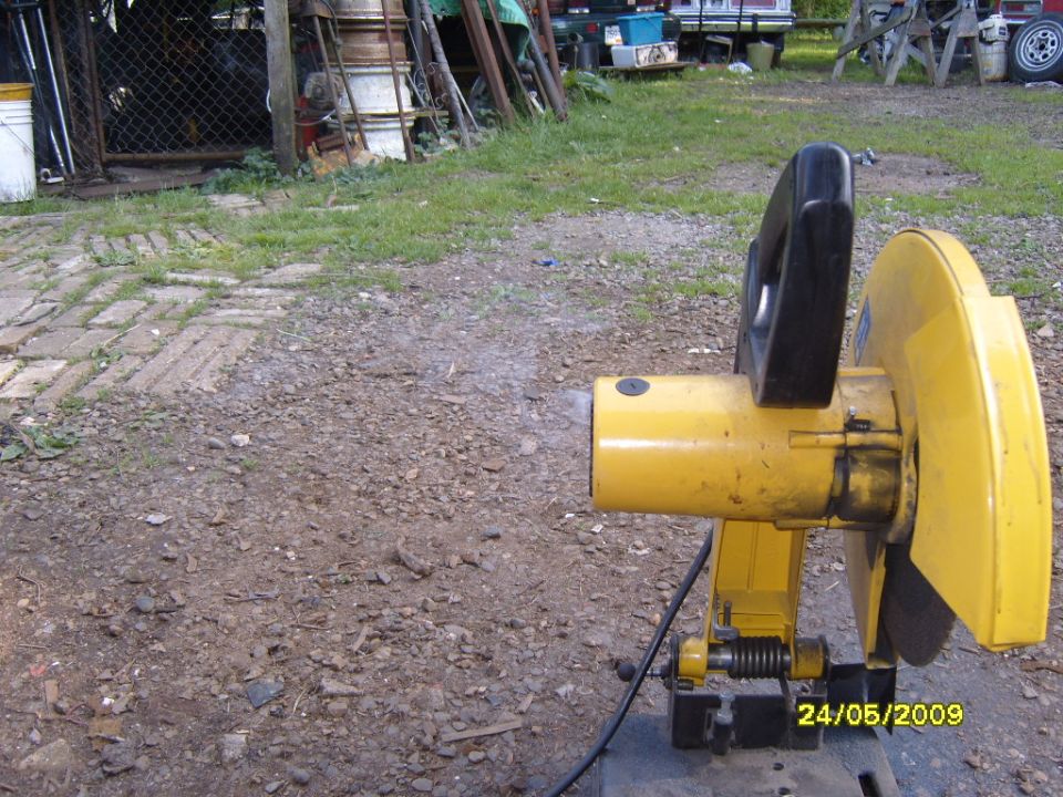 Yellow chop saw with black handle, set up outdoors on gravel. Date stamp visible