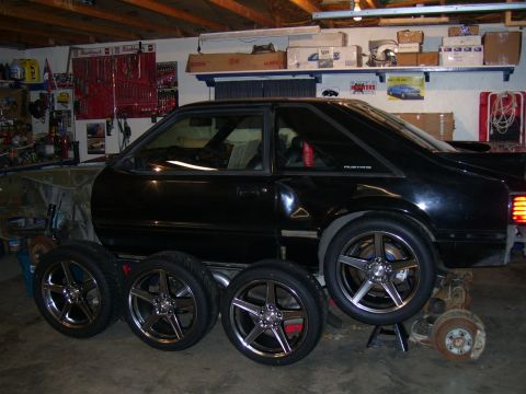 1990 Ford Mustang - Black Fox-body Ford Mustang hatchback up on stands in a cluttered home garage, with four dark-chrome five-spoke Saleen-style wheels with fr…