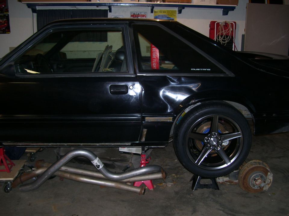 Black 1990 Ford Mustang coupe on jack stands, side view, with custom five-spoke wheels and exhaust pipes on the ground