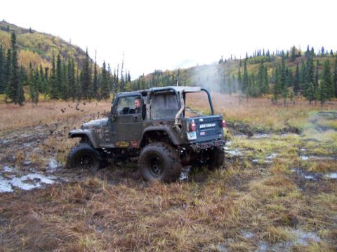 1975 Toyota Land Cruiser - Jeep Wrangler navigating a muddy off-road trail with trees and hills in the background.