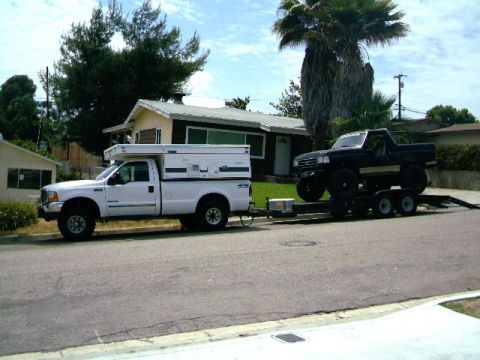 2000 Ford F-250 - A white pickup truck tows a trailer with a black truck on it down a residential street.
