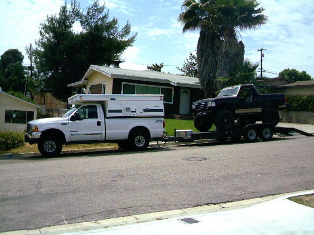 White pickup truck with a camper shell towing a trailer carrying a black lifted truck