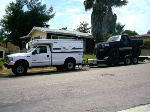 2000 Ford F-250 - A white pickup truck tows a trailer carrying a black pickup truck on a residential street.