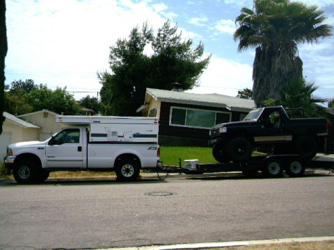 2000 Ford F-250 - A white 2000 Ford F-250 equipped with a camper tows a black truck on a trailer down a residential street.