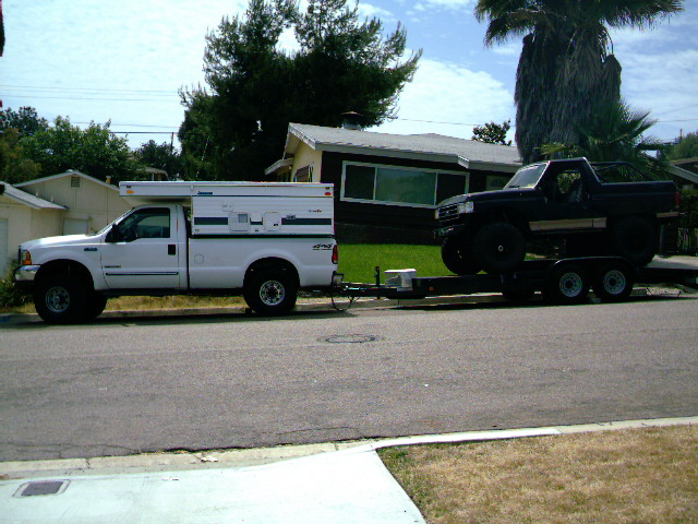 White 1997 Ford F-250 with a camper shell, towing a trailer with a black Bronco on it