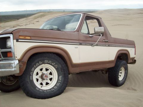 1985 Ford Bronco - This two-tone brown and white Ford Bronco is shown in a sandy, desert-like environment. The "Radial Baja" text is visible on the tire.