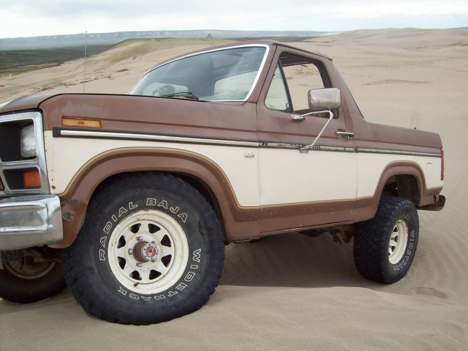 Brown and white 1985 Ford Bronco 3/4 front view on sand dunes. "Radial Baja" text visible on tire