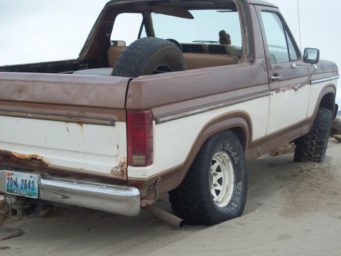 1985 Ford Bronco - Rear view of a weathered 1980s Ford Bronco with a flat tire on the beach.