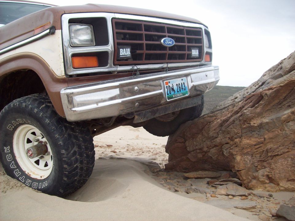 Front view of a brown and white Ford Bronco with a Baja grille, climbing over a large rock on a sandy terrain
