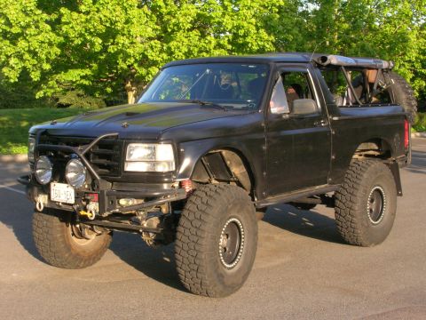 1996 Ford Bronco - Three-quarter front view of a black full-size Ford Bronco built for off-road with the roof removed and an exposed roll cage, tall mud-terra…
