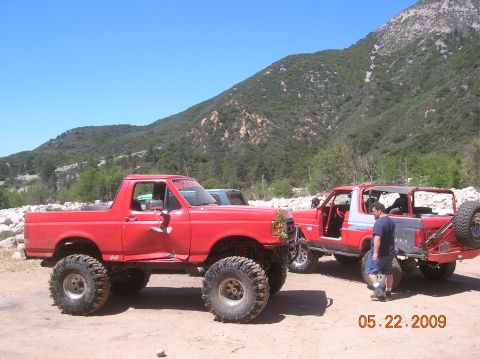 1985 Ford Bronco - Two red off-road SUVs are parked on a dirt lot with a mountainous landscape behind them under a clear blue sky.
