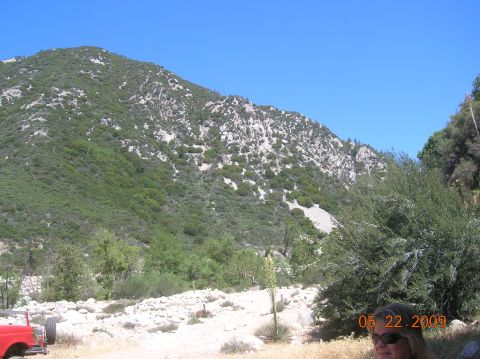 1985 Ford Bronco - A red vehicle is parked in a dry, rocky area with a large, green, tree-covered mountain under a clear blue sky.