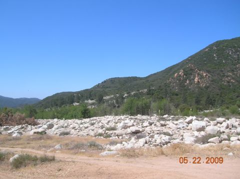 1985 Ford Bronco - A wide view of a dry, rocky landscape with a dirt path in the foreground and a tree-covered mountain in the background.