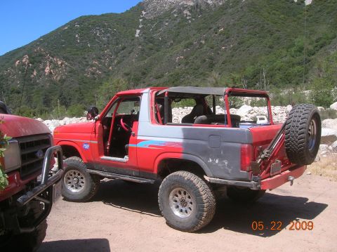 1985 Ford Bronco - Two off-road vehicles are parked on a dirt path with a mountainous backdrop.