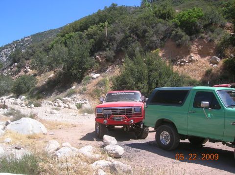 1985 Ford Bronco - Two Ford Broncos, one red and one green, are parked on a dirt road in a mountainous area.