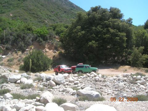 1985 Ford Bronco - Two off-road vehicles are parked in a dry creek bed with trees and mountains in the background.