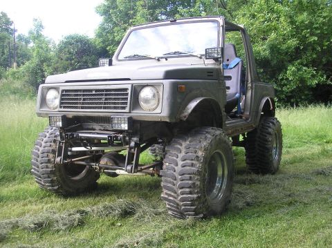 1987 Suzuki Samurai - Modified 1987 Suzuki Samurai with significant lift and large tires sits in a grassy outdoor area.