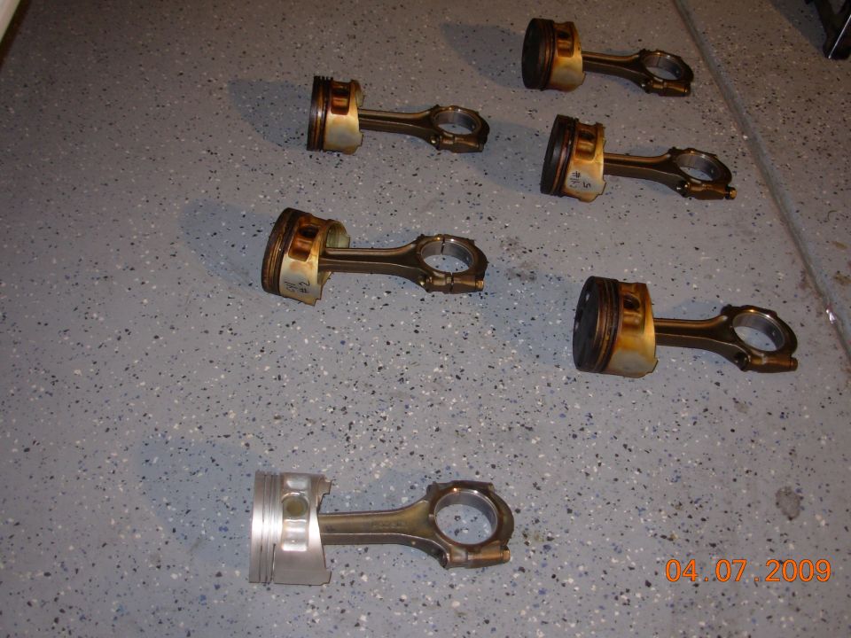 Six used pistons with connecting rods on a gray speckled floor. Some pistons show carbon buildup
