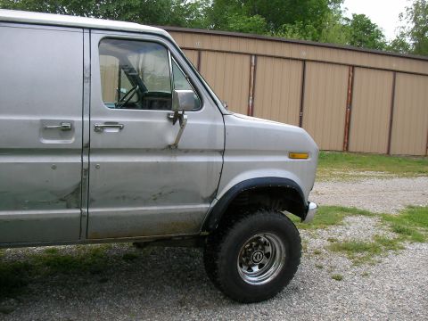 1978 Ford E250 - A silver van with a dented side panel is parked on a gravel lot in front of a tan building.