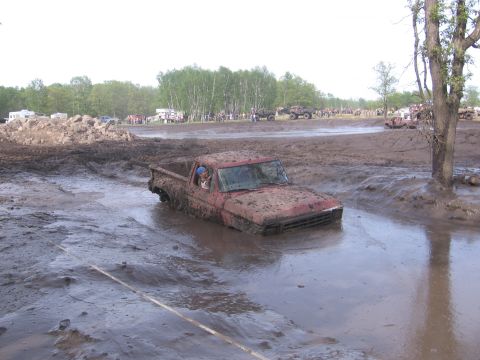 1999 Chevrolet Avalanche - A red pickup truck is submerged in mud, with spectators and other vehicles visible in the distance at a mud bog event.