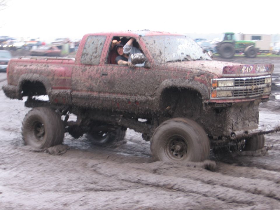 Mud-covered lifted red Chevy extended-cab pickup at a mud bog, driver waving