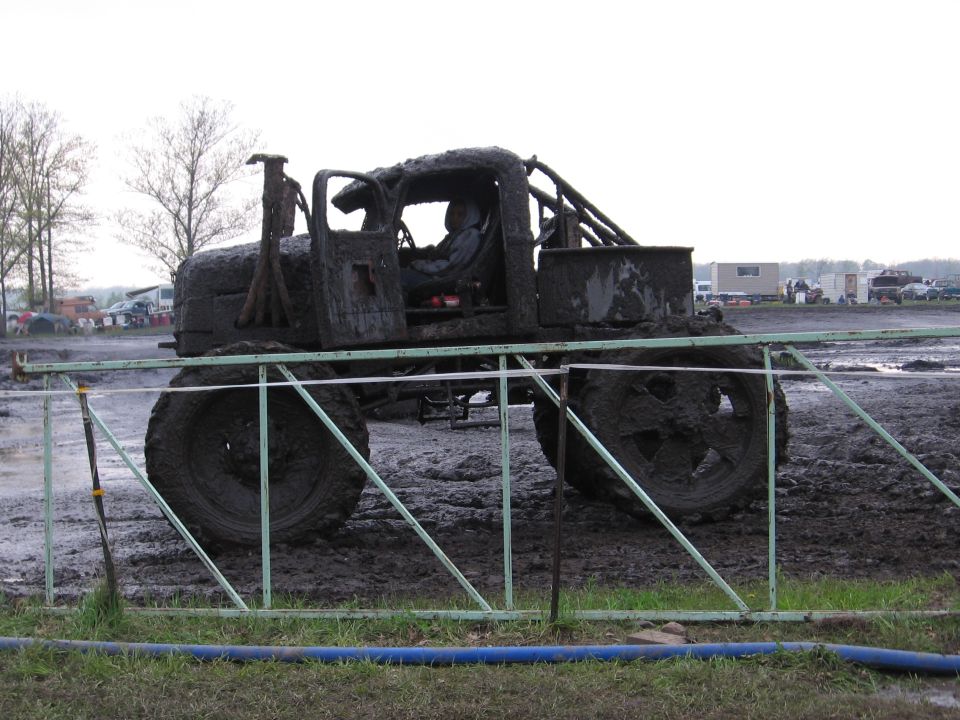 Mud-caked bog truck with roll cage and huge tires at a mud bog event, side view