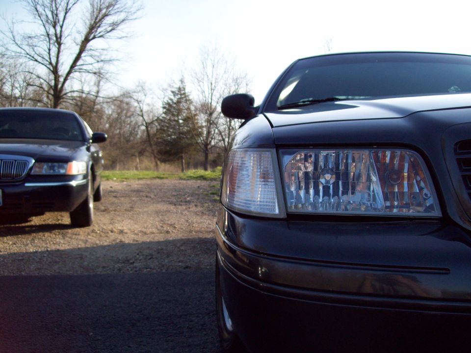 Close-up of black Crown Victoria headlight with another dark sedan parked in background on gravel lot