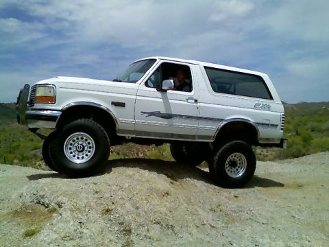 1996 Ford Bronco - This lifted 1996 Ford Bronco is shown off-roading on a dusty, rocky incline under a partly cloudy sky.
