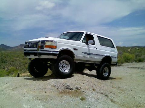 1996 Ford Bronco - A white Ford Bronco with off-road tires is parked on a dirt hill with desert vegetation in the background.
