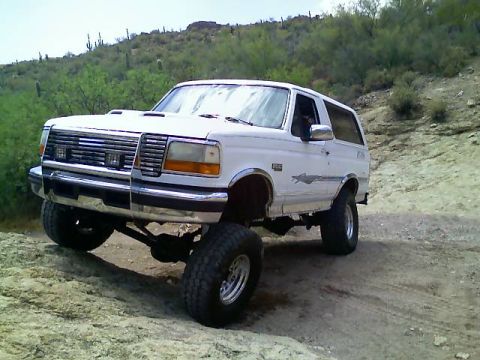 1996 Ford Bronco - White Ford Bronco with lifted suspension driving on a dirt trail in a desert landscape.