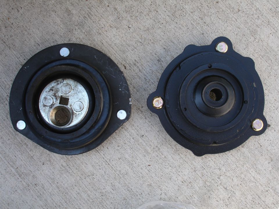 Two black strut mounts sit on a concrete surface. One has a silver metal plate with three bolts and a square hole in the center