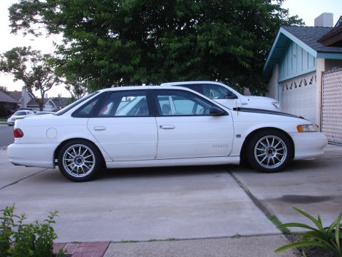 1993 Ford Taurus - A white 1993 Ford Taurus sedan is parked in a driveway, showing its side profile and a glimpse of its interior racing cage.
