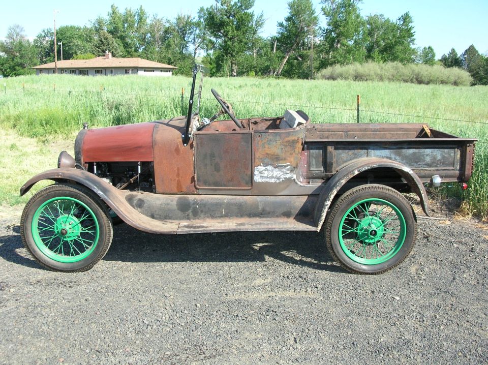 Rusty pickup truck with green wheels, red hood, and a wooden bed. It's parked on gravel with a house and field in the background