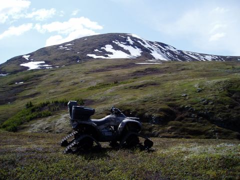 2000 Ford E350 - An all-terrain vehicle equipped with tracks sits on a grassy slope below a snow-dusted mountain.