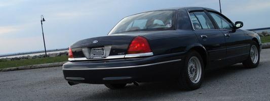 Black 2000 Ford Crown Victoria sedan, rear 3/4 view, parked on asphalt near water